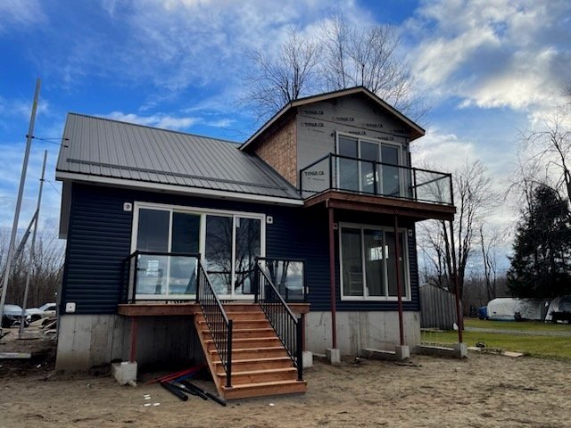 New two-story house with a metal roof, black siding, and large windows, featuring balconies and front stairs, surrounded by trees and open sky.