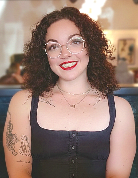A woman with curly brown hair, glasses, red lipstick, and a black sleeveless top, smiling in a cafe setting.