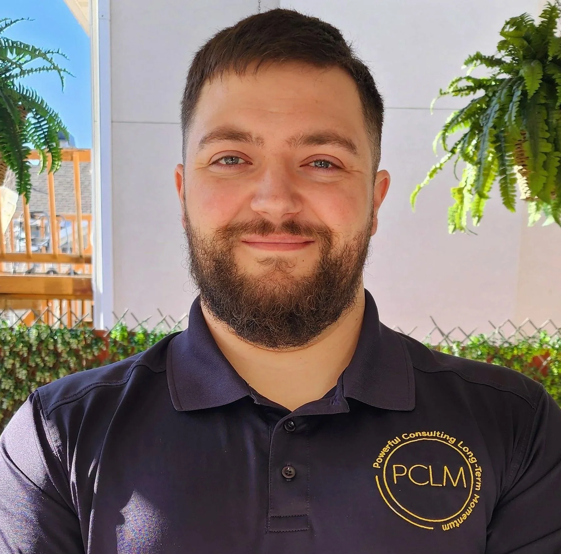 A smiling man with a beard and short brown hair wearing a black polo shirt with a logo on the chest that reads 'PCLM' and 'Powerful Consulting Long-Term Momentum.' He is standing outdoors in front of a fence, with green plants and a building in the background.