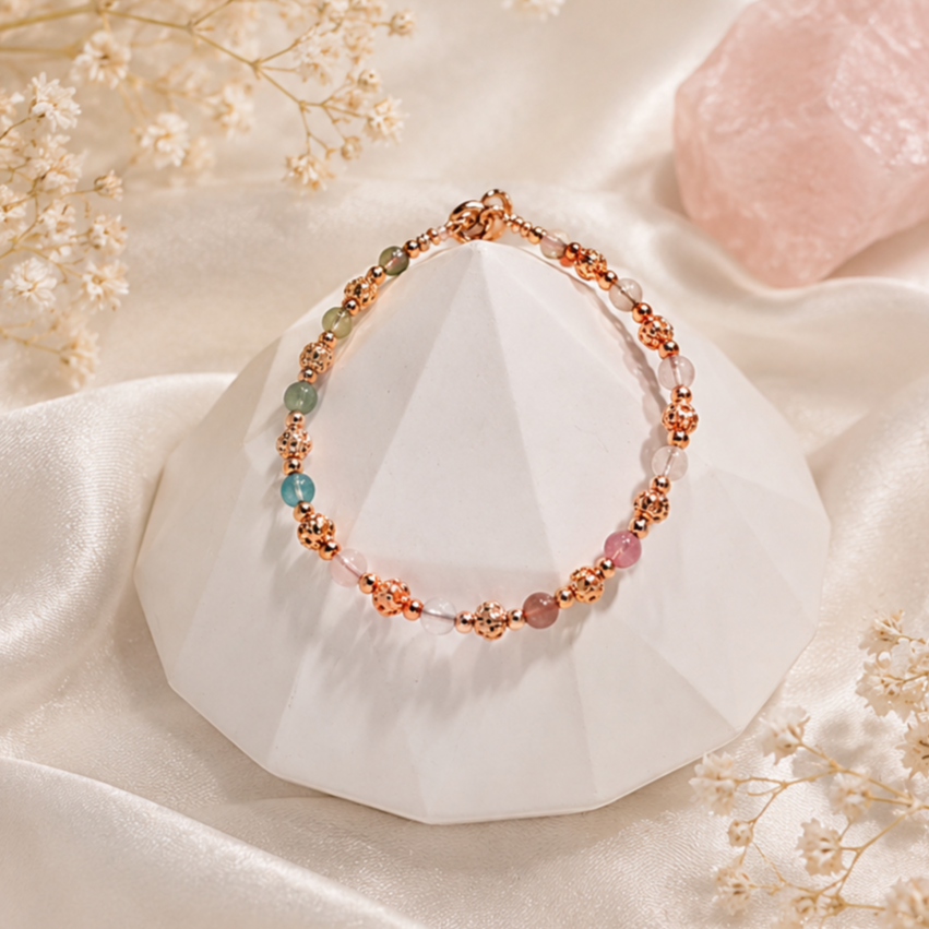 Elegant multicolored beaded bracelet with rose gold accents displayed on a white geometric stand, surrounded by soft silk and dried white flowers with a pink salt rock in the background.