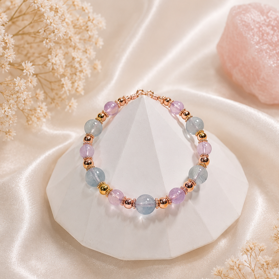Beaded bracelet with pastel purple, blue, and gold beads on a white geometric display, surrounded by light pink flowers and a pink mineral on cream satin fabric.