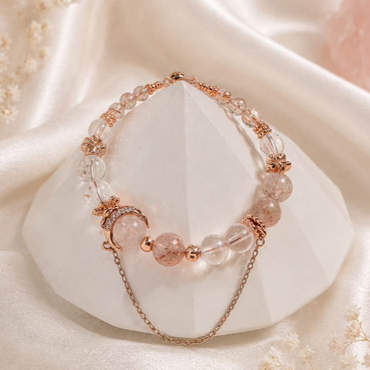 A necklace with pink and clear beads, gold accents, and a moon charm draped over a white geometric stone on satin fabric.