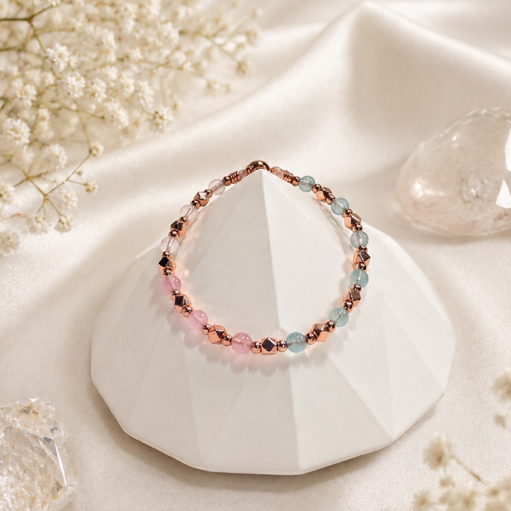 A beaded bracelet with pink, blue, and metallic beads displayed on a white geometric ceramic piece, with small flowers and crystals in the background.