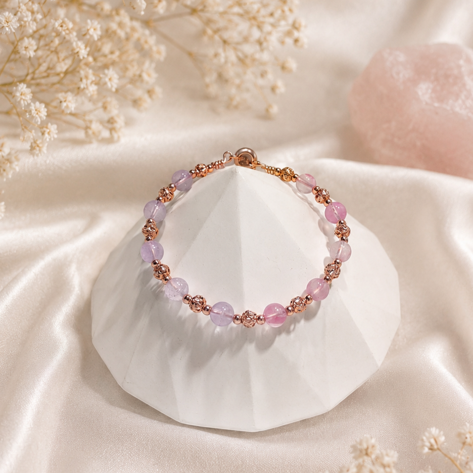 Pink and purple beaded bracelet with rose gold accents on a white geometric display, surrounded by cream-colored flowers and a pink mineral rock.