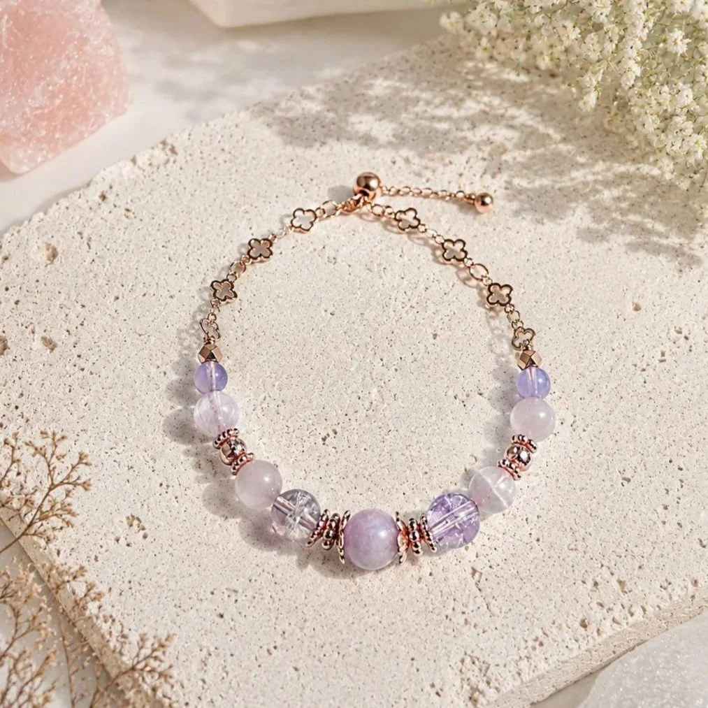 A delicate bracelet with purple and clear beads, gold accents, and a floral chain, displayed on a sandy textured surface with dried flowers in the background.