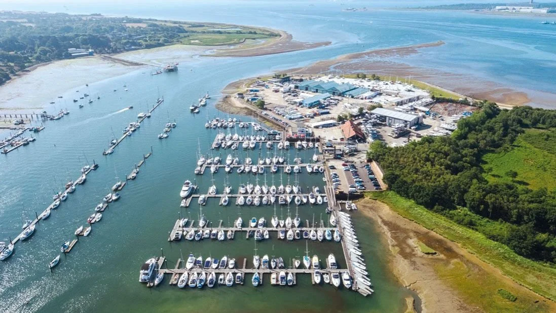 Aerial view of a marina on Hamble River with numerous boats docked in rows, adjacent to a facility with parking and surrounding green landscape.