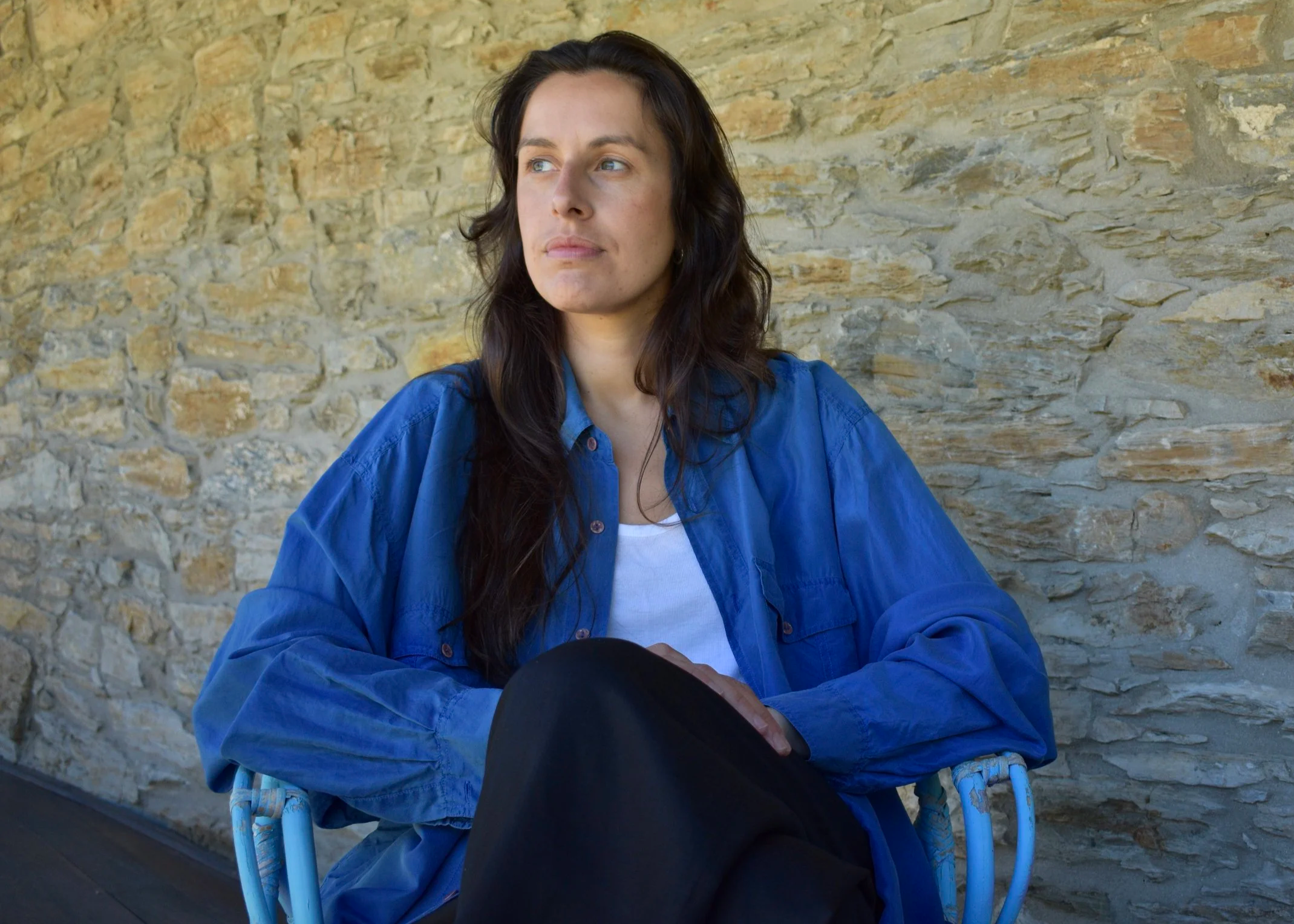 Mujer con cabello largo y oscuro, vestida con camiseta blanca y una chaqueta azul, sentada en una silla, frente a una pared de piedra