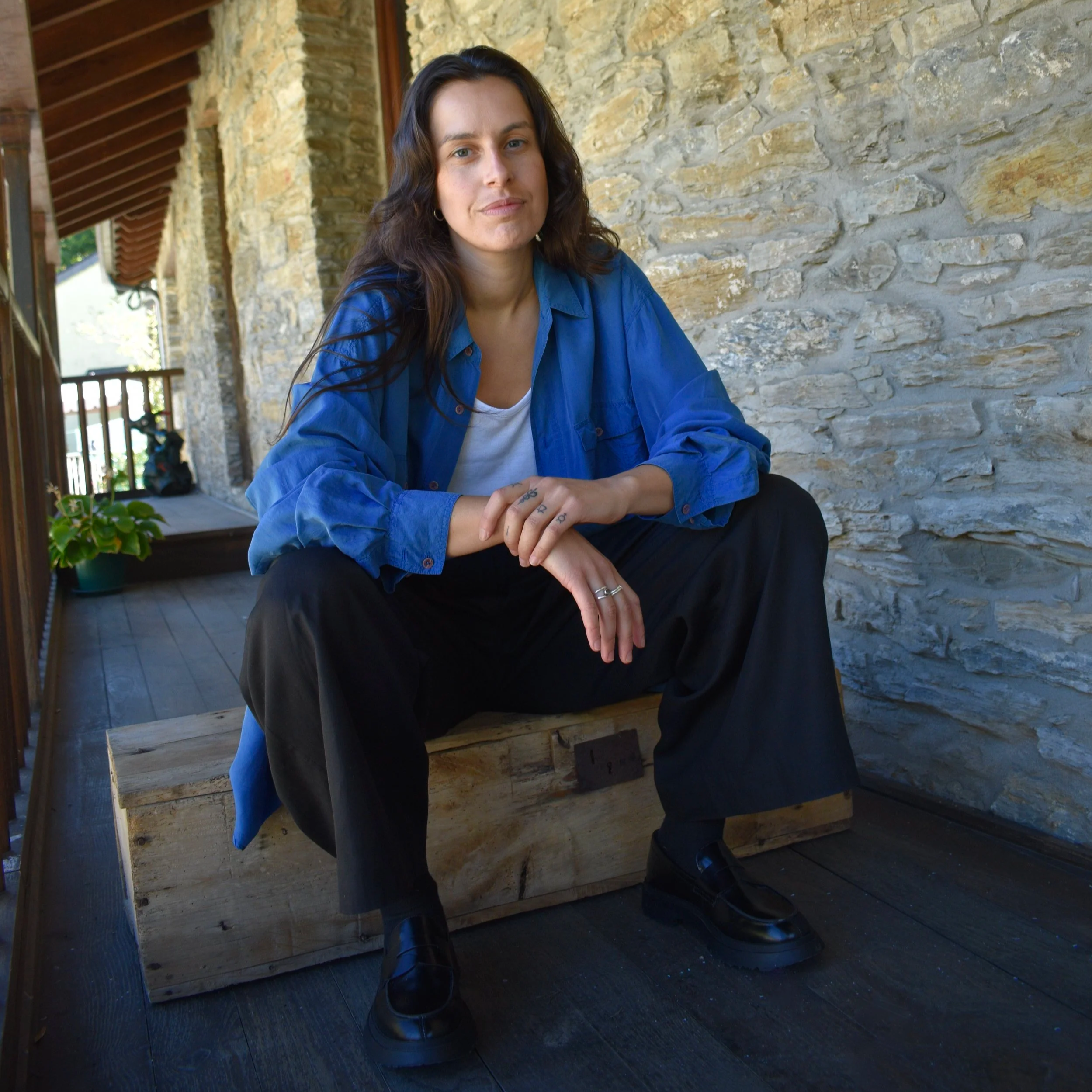 Joven mujer con camiseta blanca, chaqueta azul y pantalones negros, sentada en un banco de madera en un balcón de piedra, con plantas y estructura de madera en segundo plano.