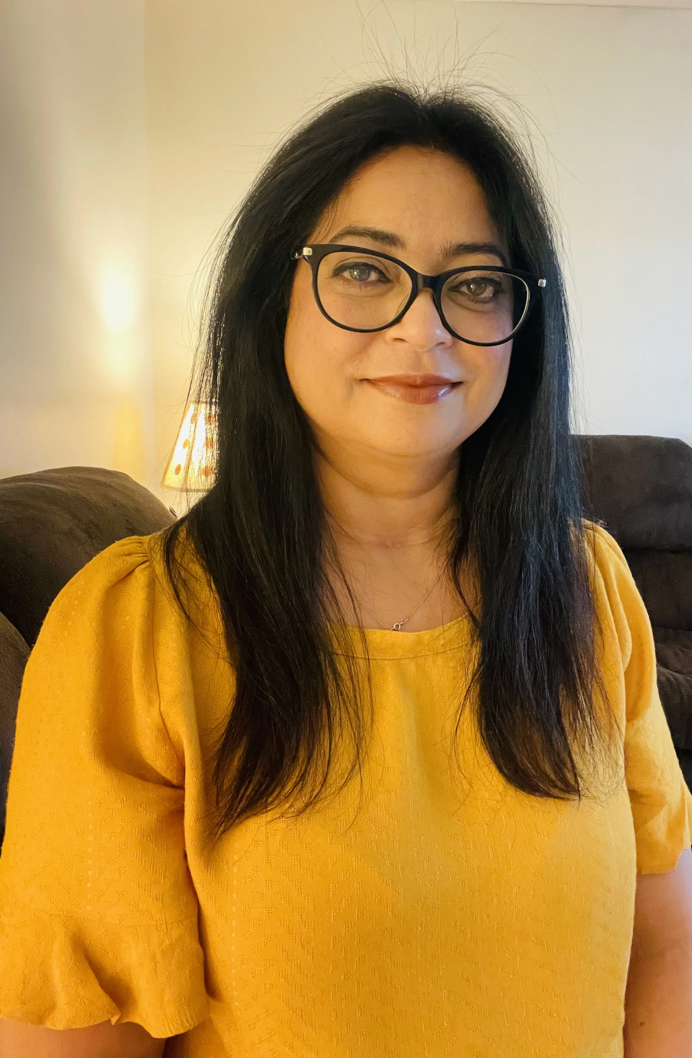 A woman with long dark hair, glasses, and a yellow blouse sitting on a couch in a cozy room with a lamp in the background.