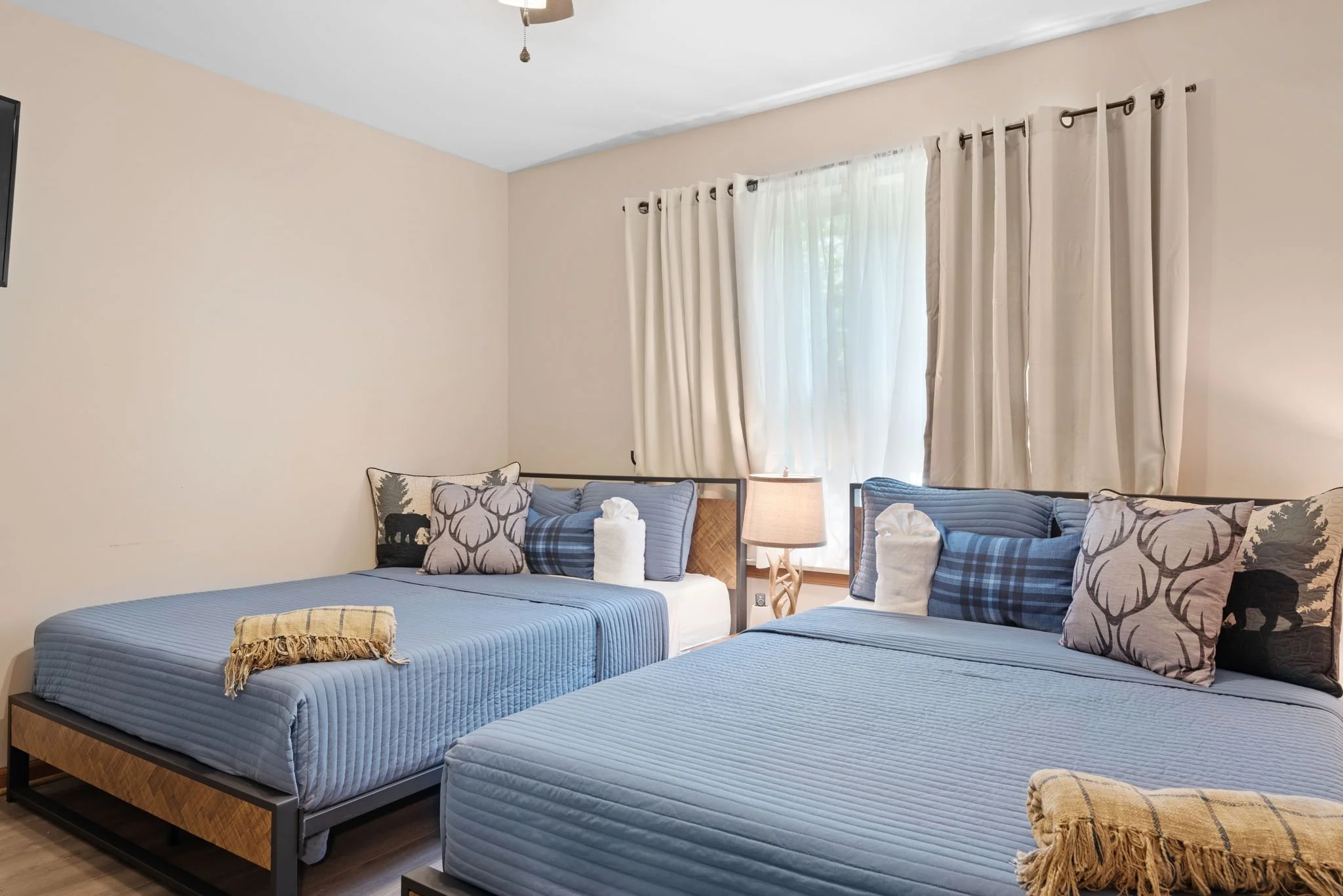 A bedroom with three beds, each made with blue striped mattresses and decorated with various pillows, including one with black bears, one with tree designs, and plaid patterns. A side table with a beige lamp is positioned between two of the beds. The room has a window with cream-colored curtains and sheer white curtains, letting in natural light.