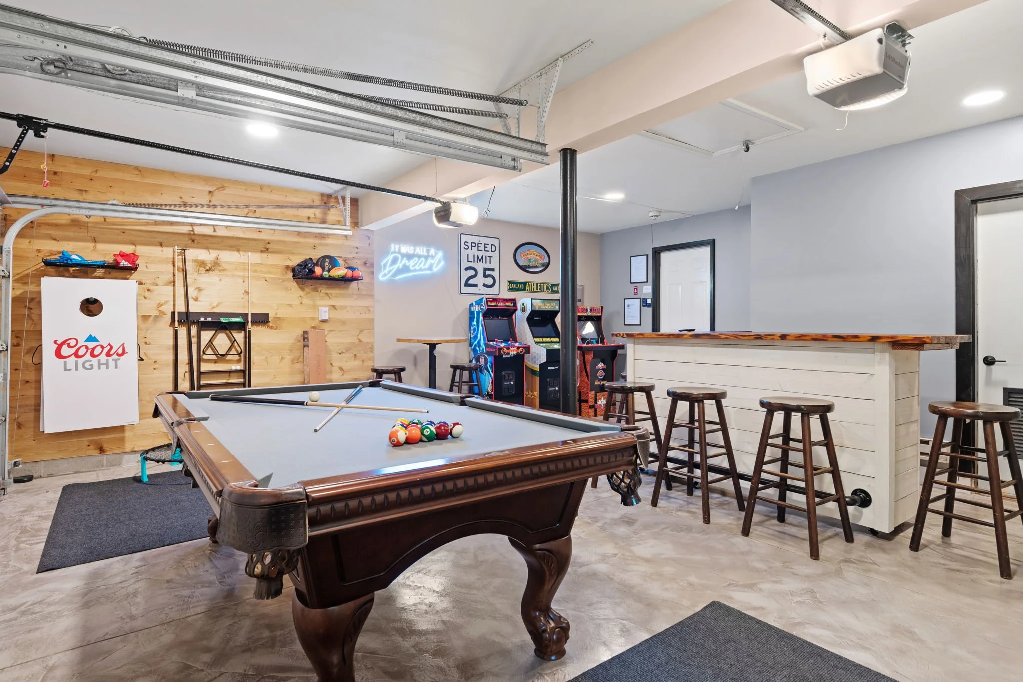 Indoor recreational space with pool table, arcade machines, bar area, stools, and sports-themed wall decor.
