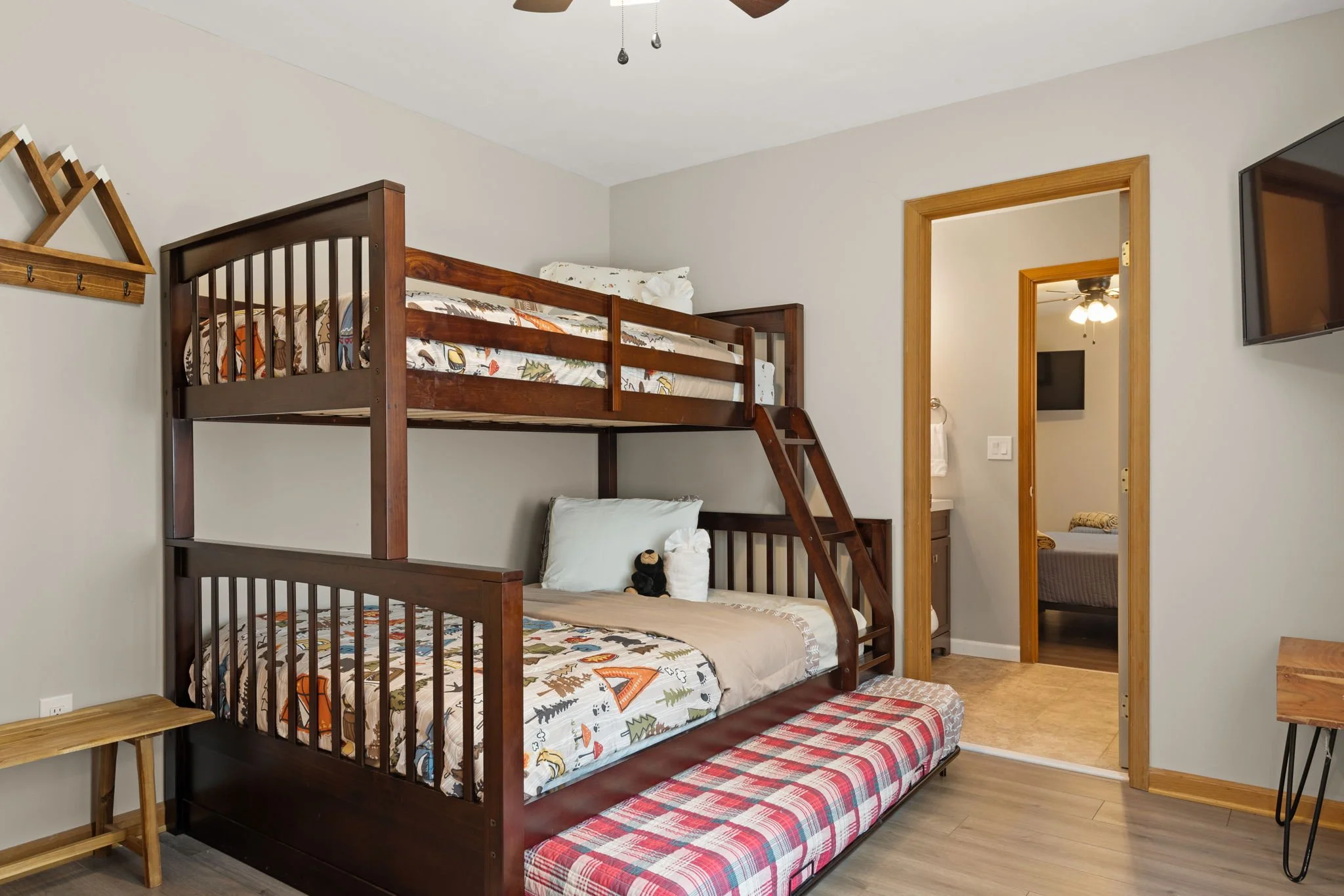 A bedroom with a wooden bunk bed featuring a triple twin setup and a ladder. The bed has patterned sheets with outdoorsy designs. There is a small wooden side table, a plaid-patterned bench, and a flat-screen TV on the wall. The room has wooden door frames and light-colored walls with a ceiling fan.