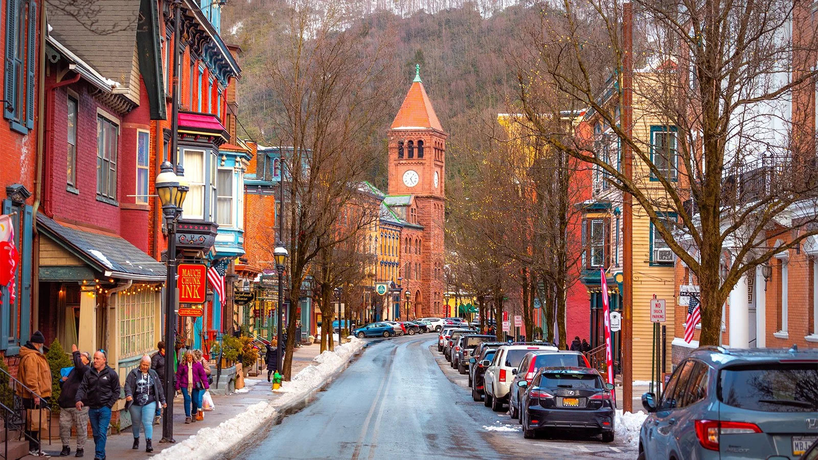 Colorful historic buildings along a street with parked cars, people walking, leafless trees, and a clock tower in the background on a winter day.