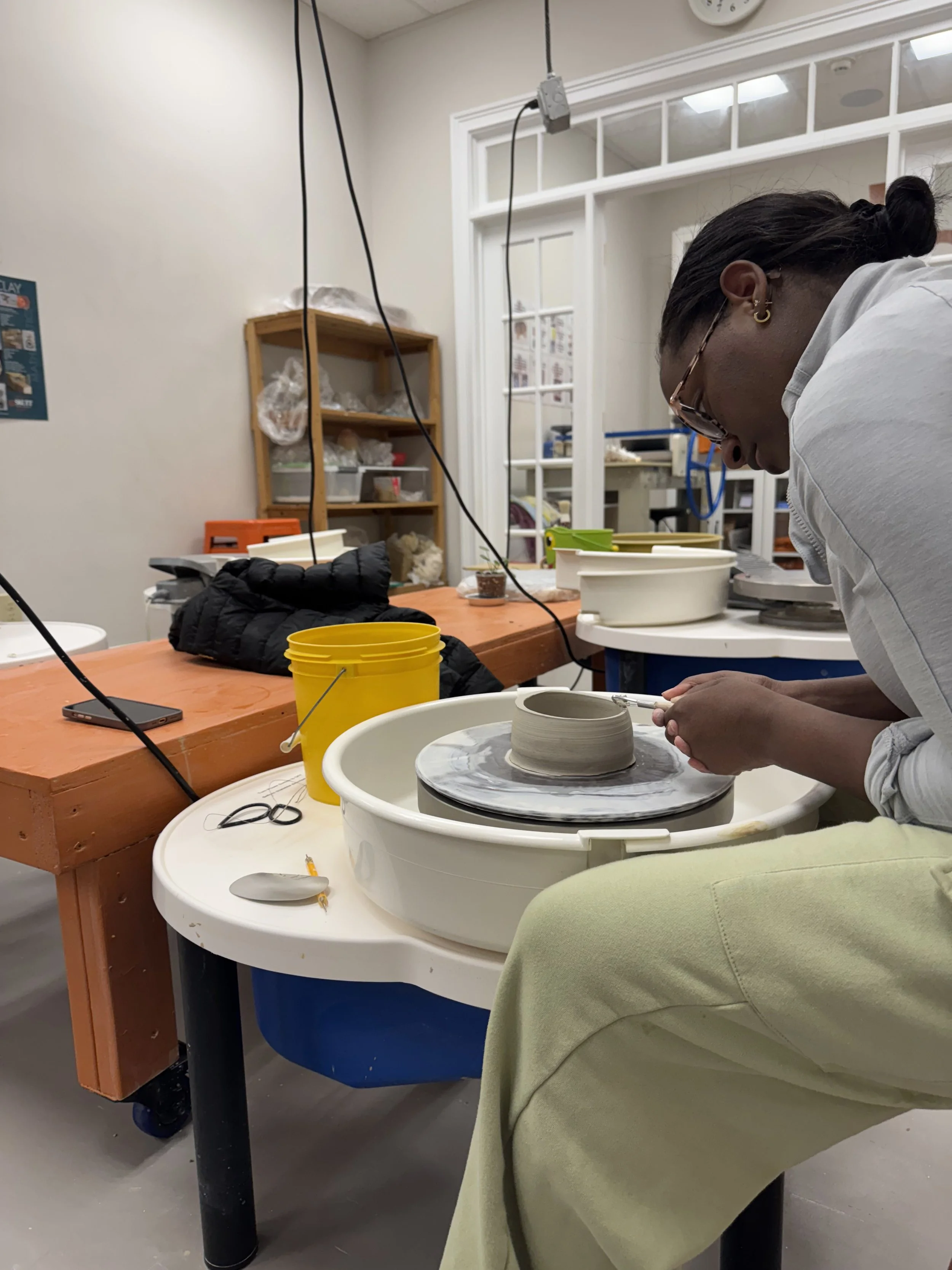 Person shaping pottery on a wheel in a ceramics studio with tools and supplies nearby.