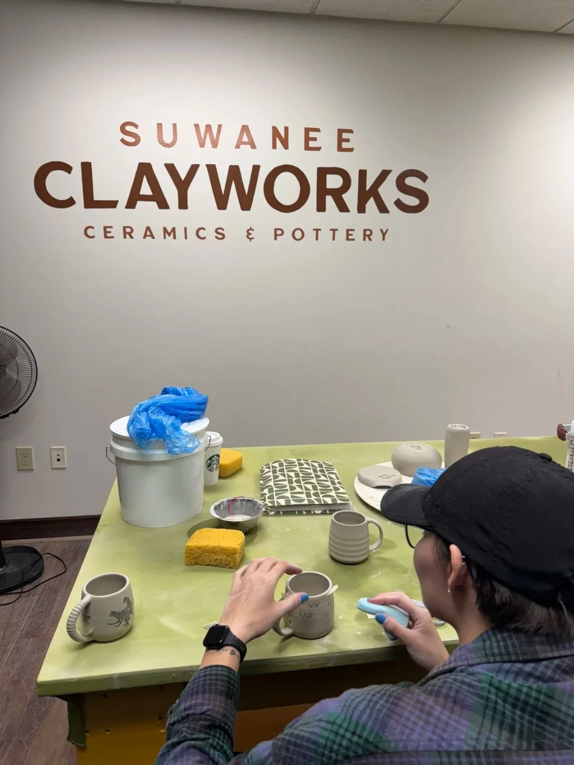 A person with short hair wearing glasses, a black cap, and a plaid shirt is working on pottery at a table in a ceramics studio called Suwanee Clayworks. The studio has a sign on the wall that reads 'Suwanee Clayworks Ceramics & Pottery.' The table is filled with various pottery items, sponges, and tools.