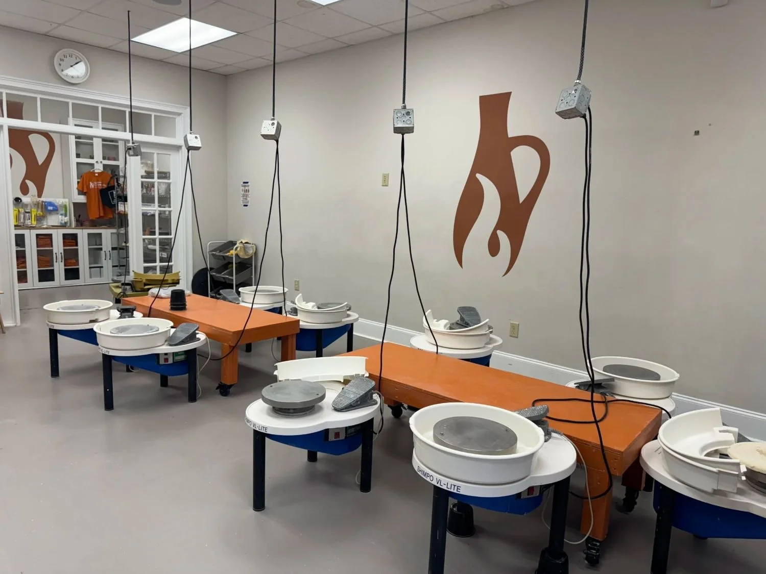 Suwanee Clayworks pottery studio with several pottery wheels and tools, decorated with large orange and brown pottery-shaped wall art.