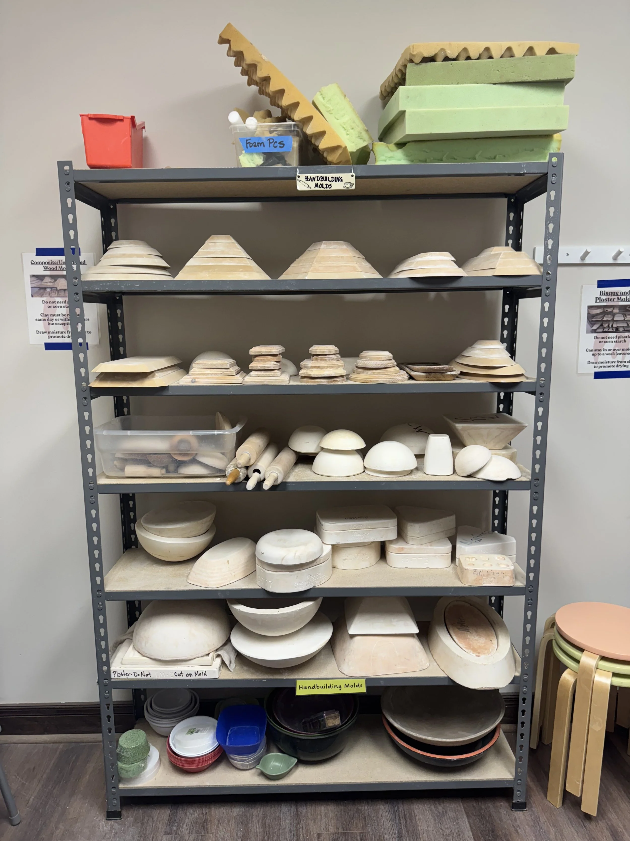 Shelves holding unpainted ceramic and plaster handbuilding molds and bumpers, with foam and green sponges on top, and stacked colored bowls and plates on the bottom.