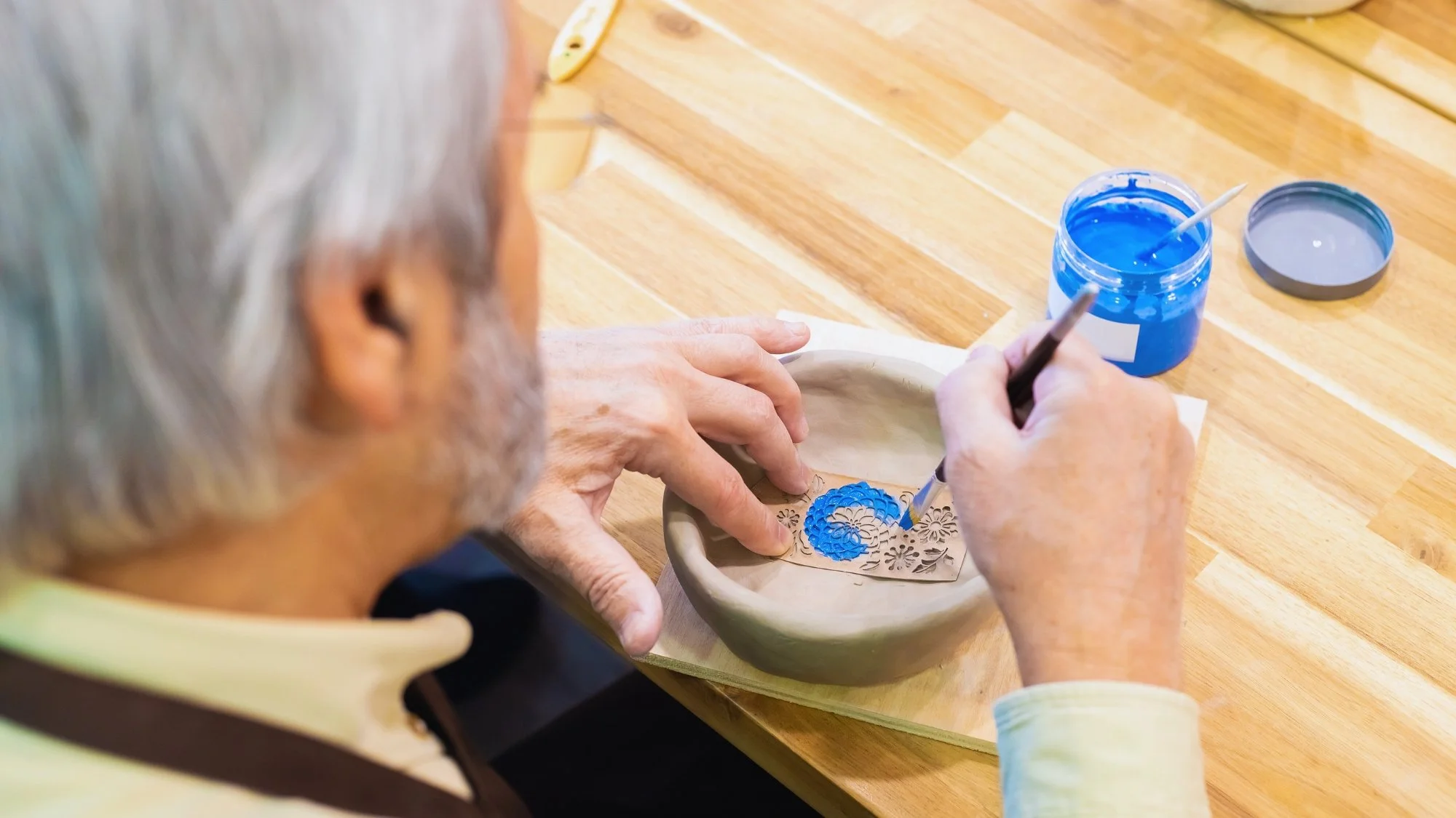 A person with gray hair is painting a blue design on a ceramic plate while sitting at a wooden table, using a small brush and a palette of blue paint.