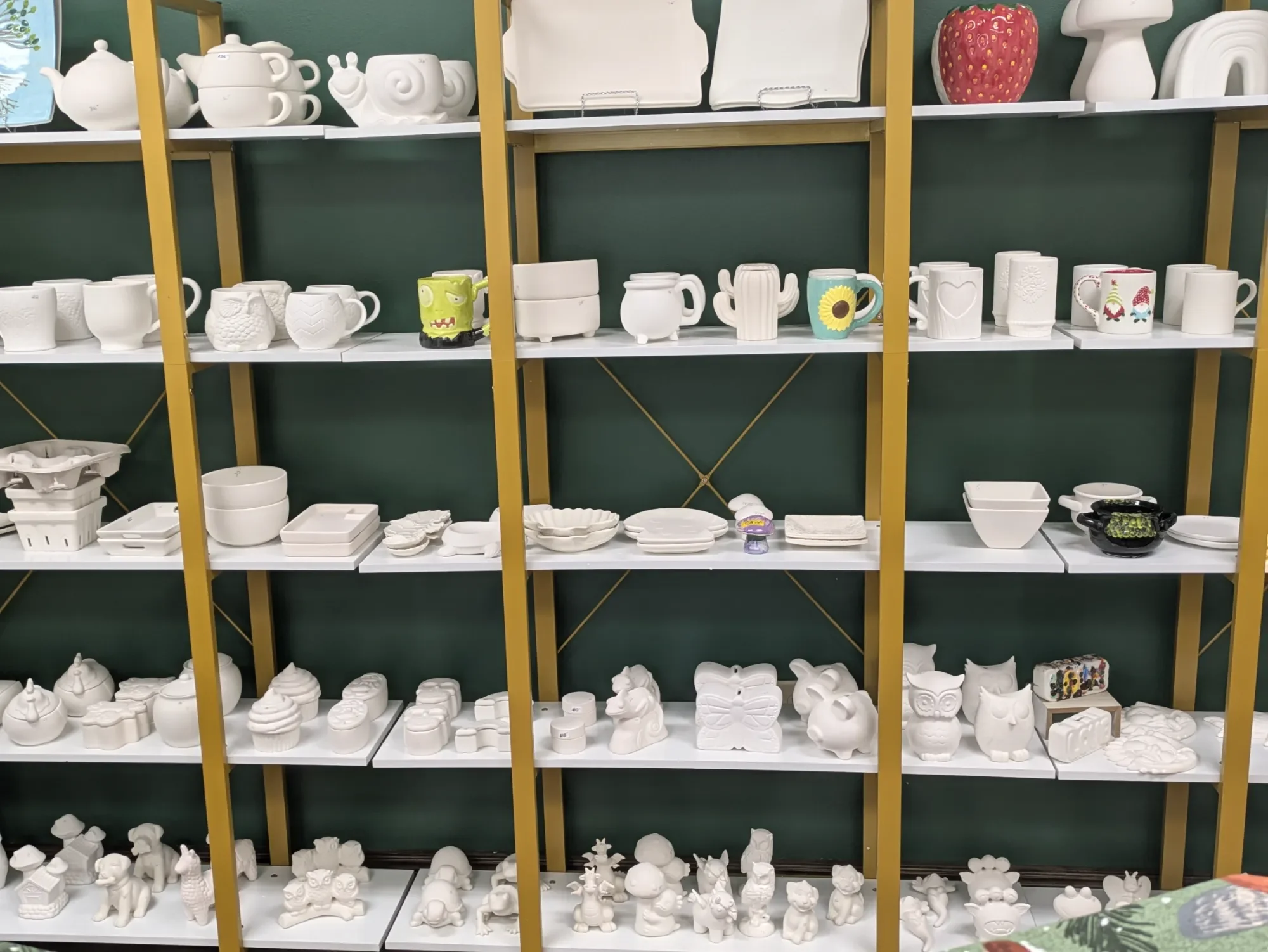 Shelves filled with various ceramic and porcelain figurines, mugs, bowls, and decorative items in white, with some colored pieces, all ready for painting.