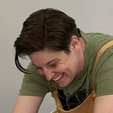 Marissa Loose working at the pottery wheel, smiling and looking down, wearing a green shirt and apron.