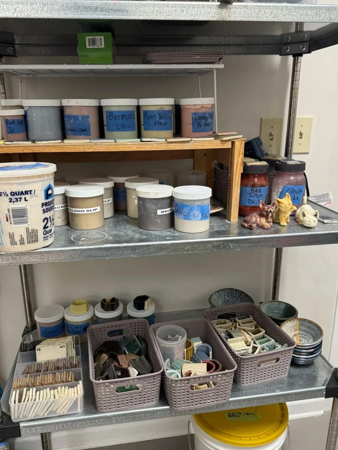 Shelf with various containers and jars holding pottery glaze supplies such as pigments, paints, and brushes, in a storage or workshop setting.