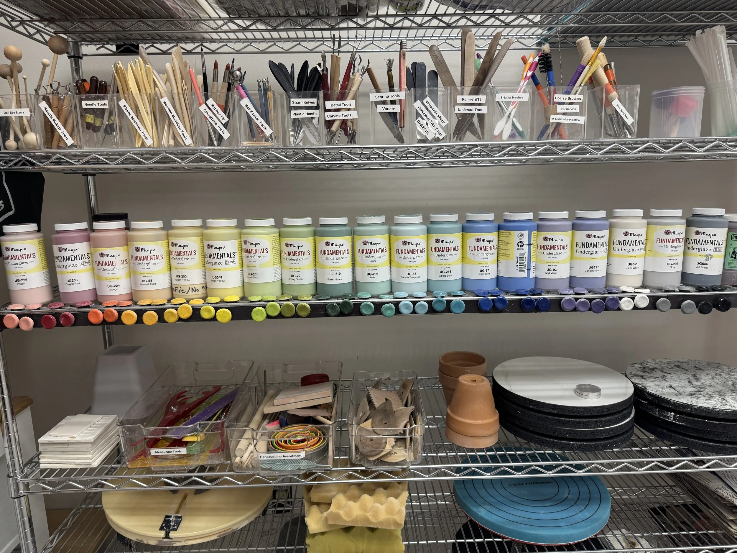 Shelving unit with various art supplies including bottles of acrylic paint, paintbrushes, carving tools, plastic containers with different tools, clay pots, circular slabs, cutting mats, and other artistic tools.