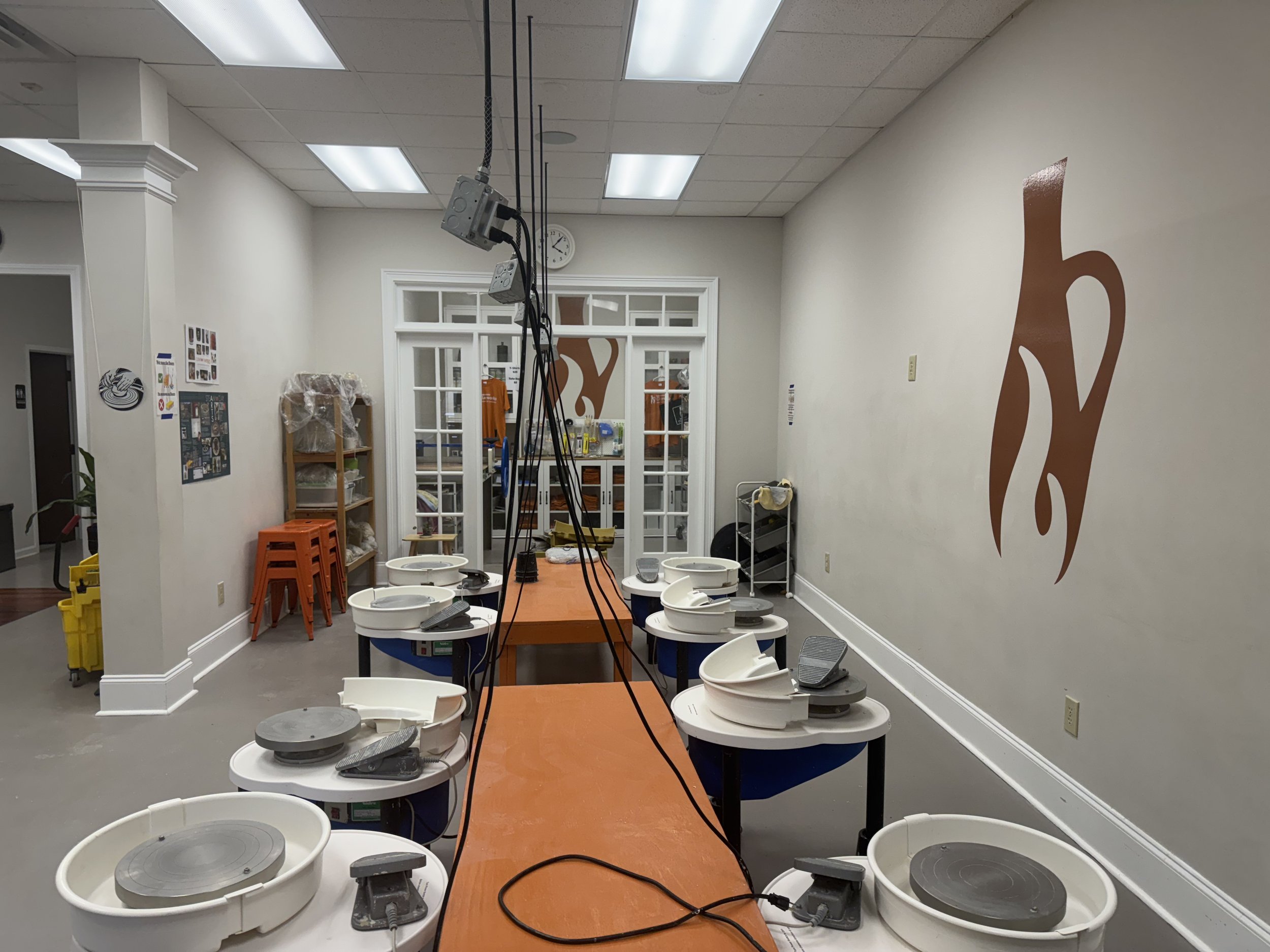 Indoor pottery studio with several pottery wheels arranged on black tables. A long orange work table runs down the center. Shelves with supplies, a wall clock, and an orange icon of a pottery vessel are visible.