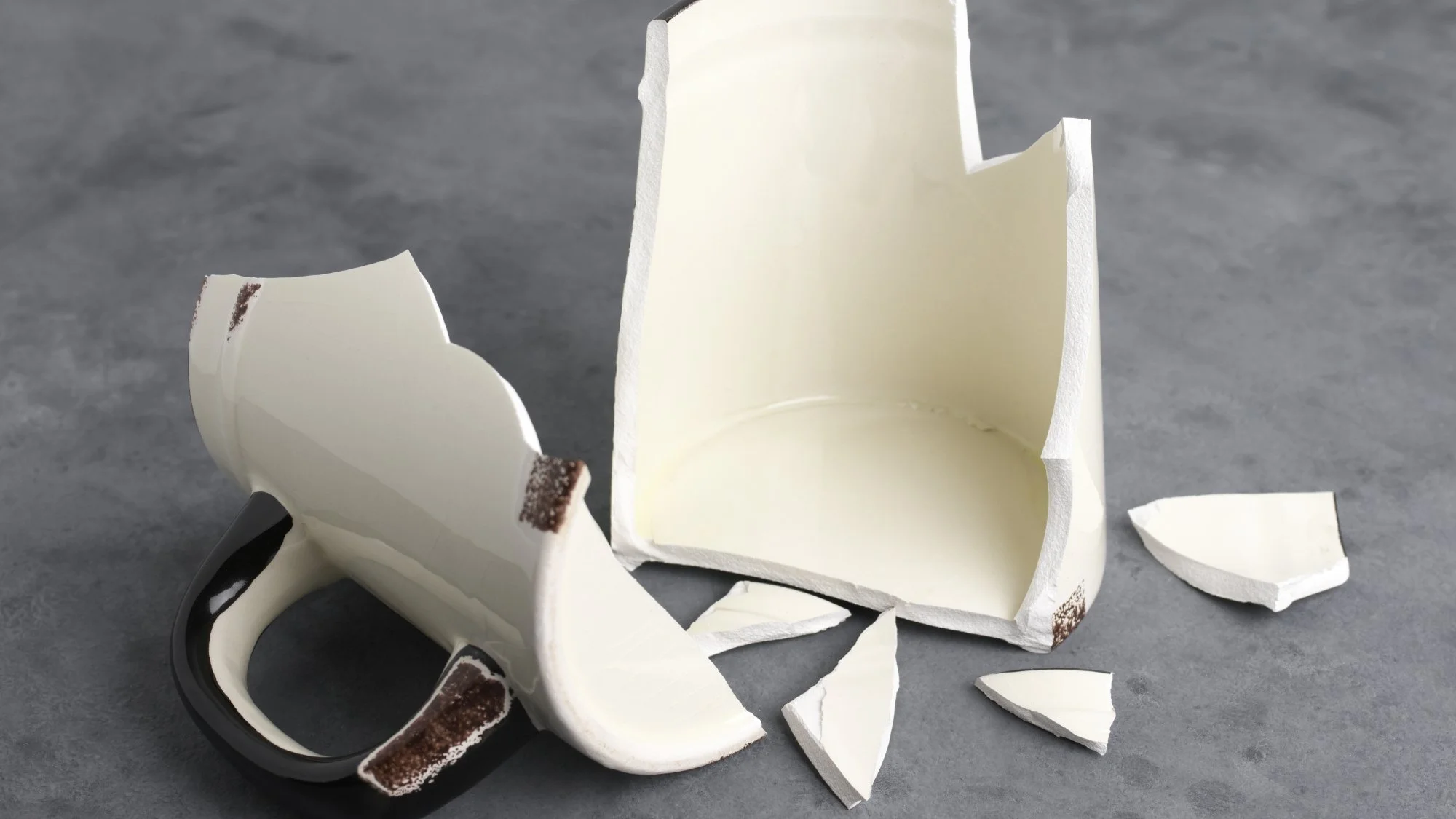 Broken ceramic mug on gray surface with scattered shards.