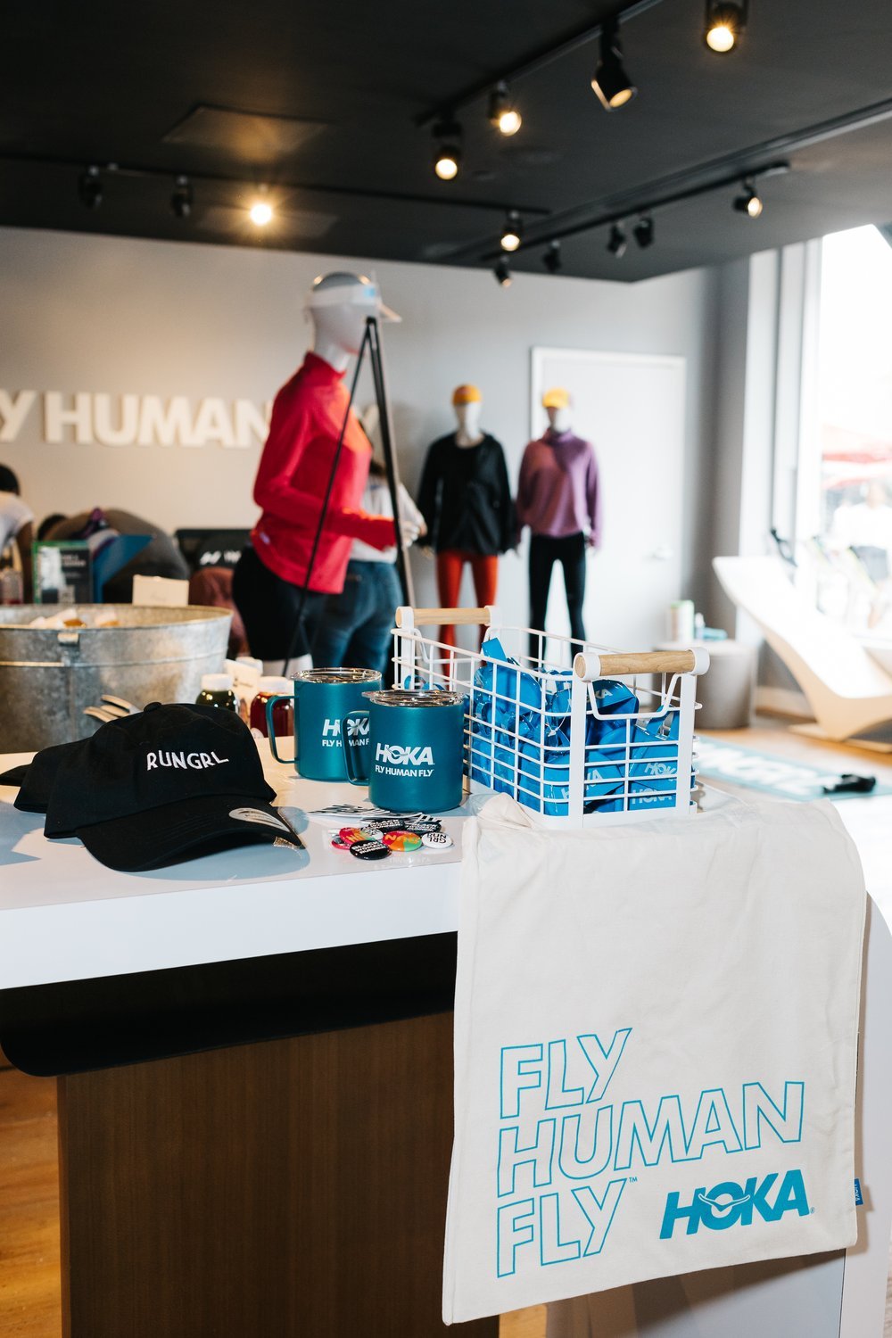 Display table with HOKA merchandise including mugs, caps, and buttons, with mannequins dressed in athletic wear in the background.