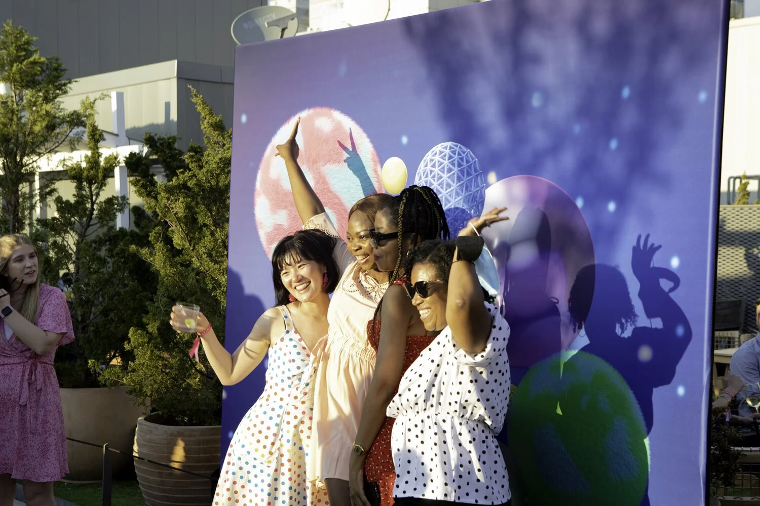 Four women at an outdoor event posing for a photo against a colorful, futuristic backdrop with geometric shapes, with other people and trees in the background.