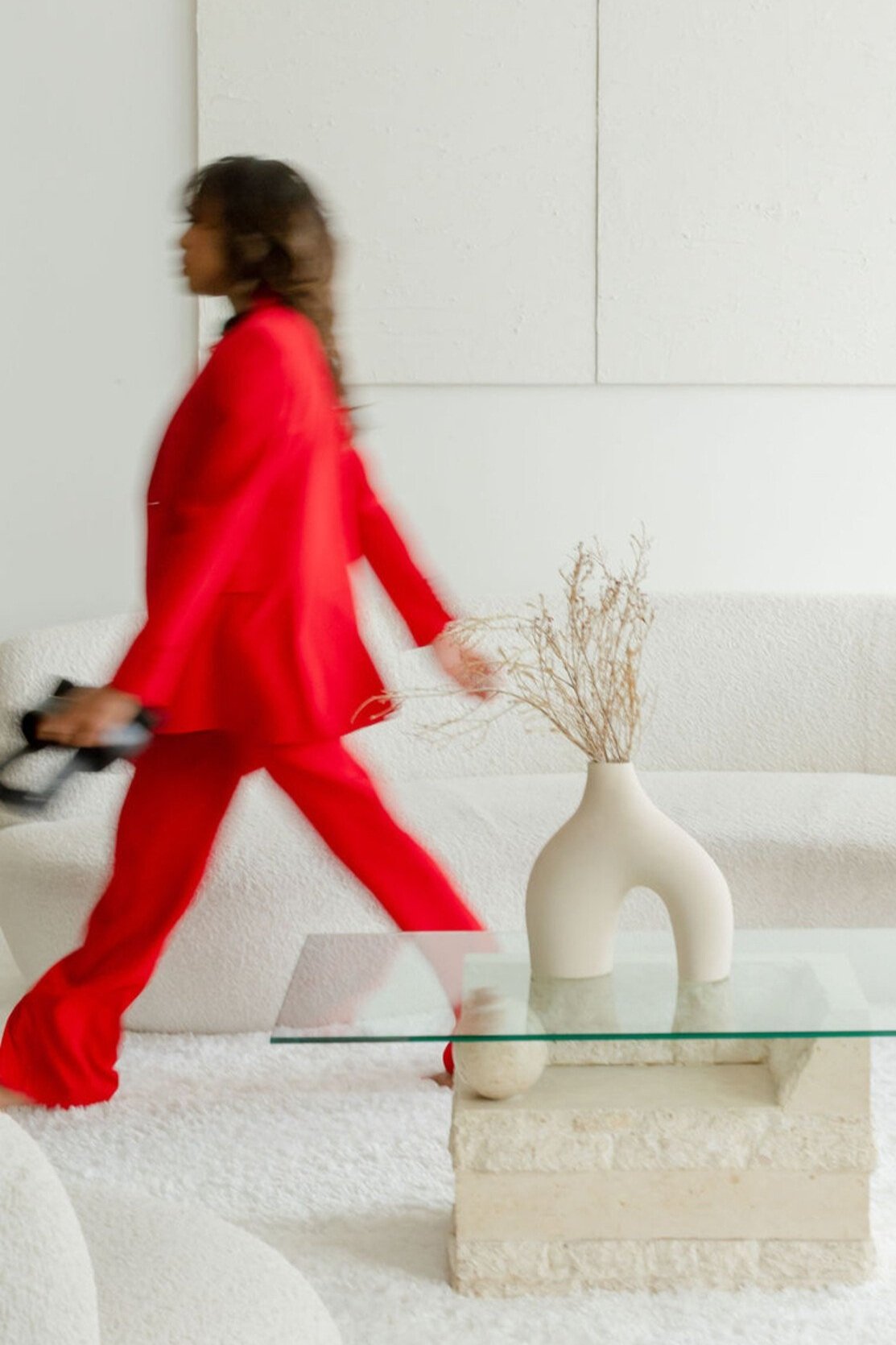 A woman dressed in a red suit walking past a white sofa and a glass coffee table with a beige vase containing dried branches