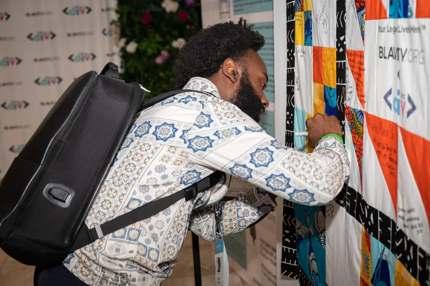 A man with a backpack signing a colorful quilt at an event.