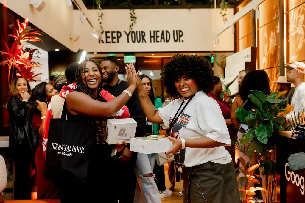 Two women smiling and high-fiving at an indoor event, surrounded by people, with a sign that says "Keep Your Head Up" and a Google branding tote bag.