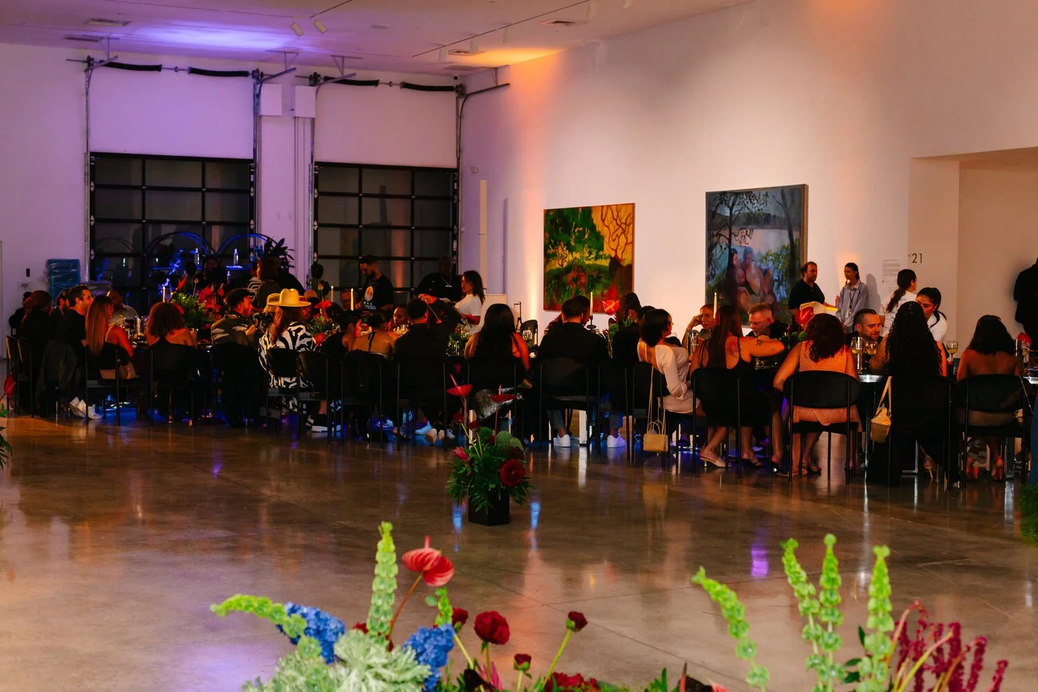 A large indoor gathering with people seated at long tables, decorated with flowers, in a well-lit art gallery with paintings on the walls.