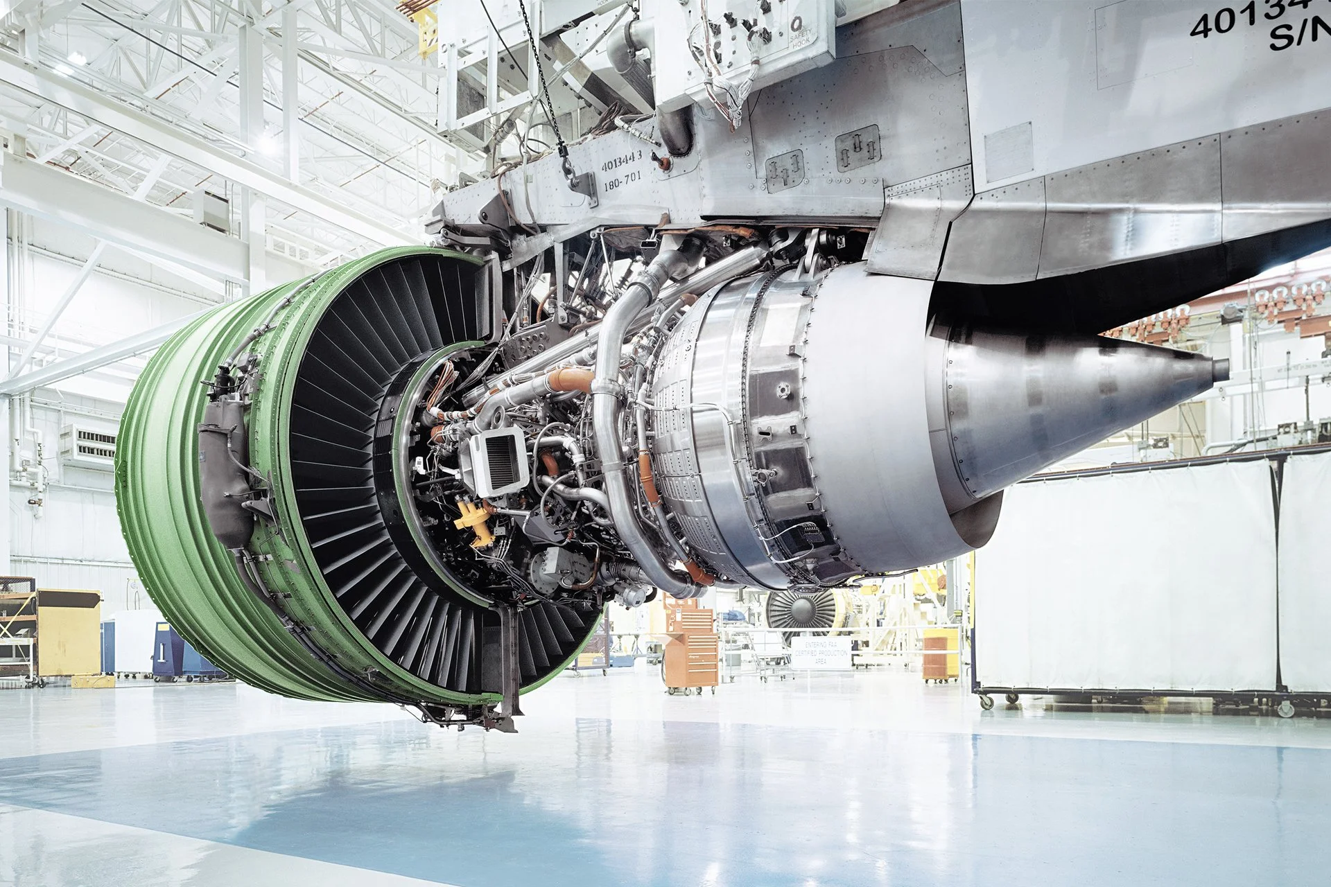 Close-up view of a large aircraft jet engine inside a spacious, well-lit hangar, with various industrial and maintenance equipment around.