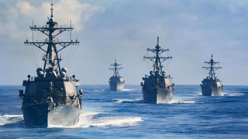 Four navy ships sailing in formation across the ocean under a partly cloudy sky.