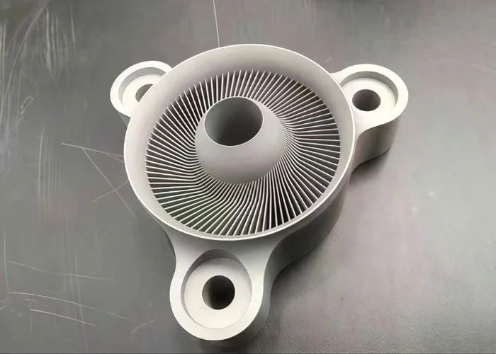 A gray plastic or metal component with a circular, vent-like structure and three mounting holes.