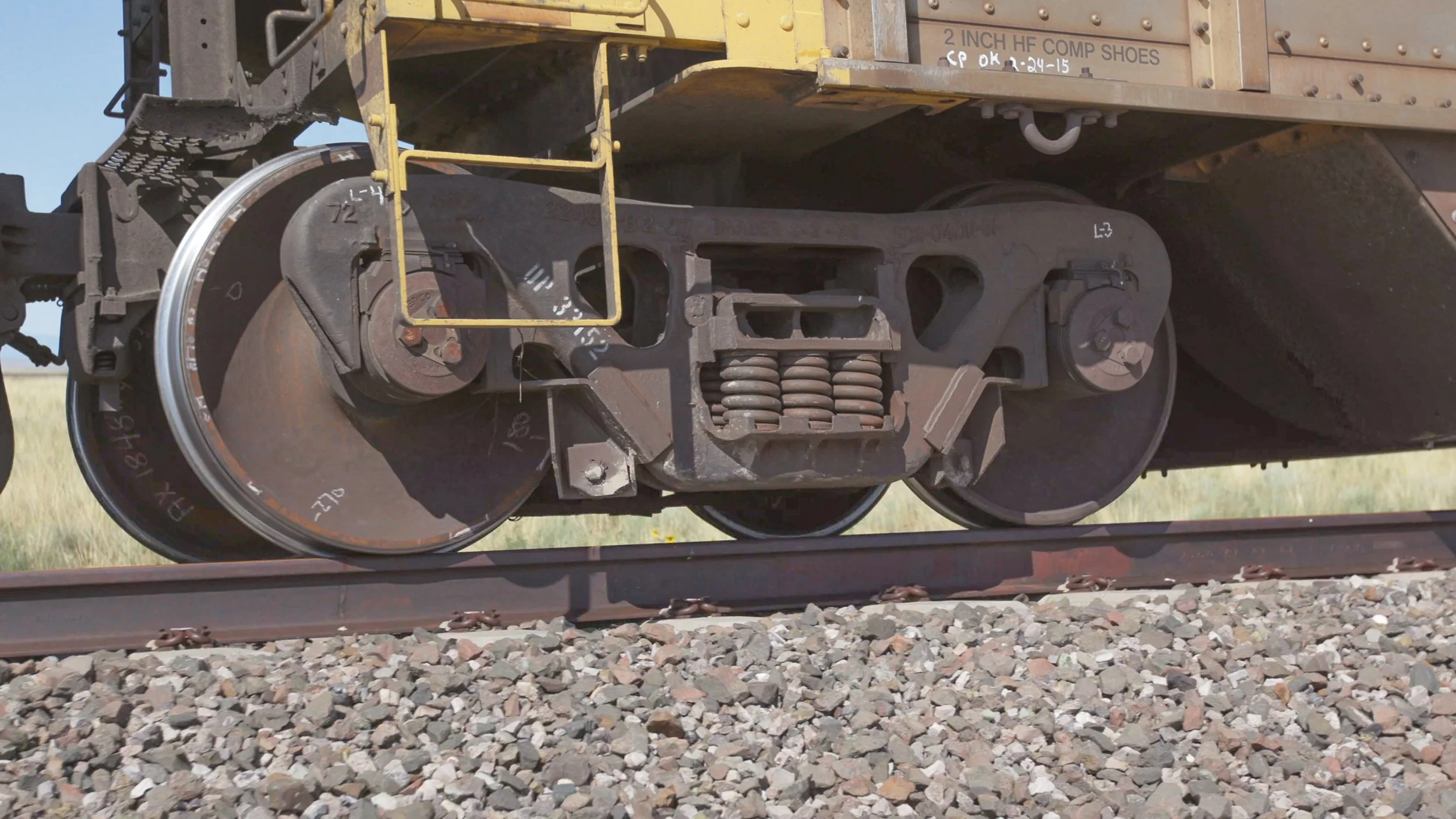 Close-up of a railway wheel and coupling mechanism on a train, with gravel on the tracks in the foreground.