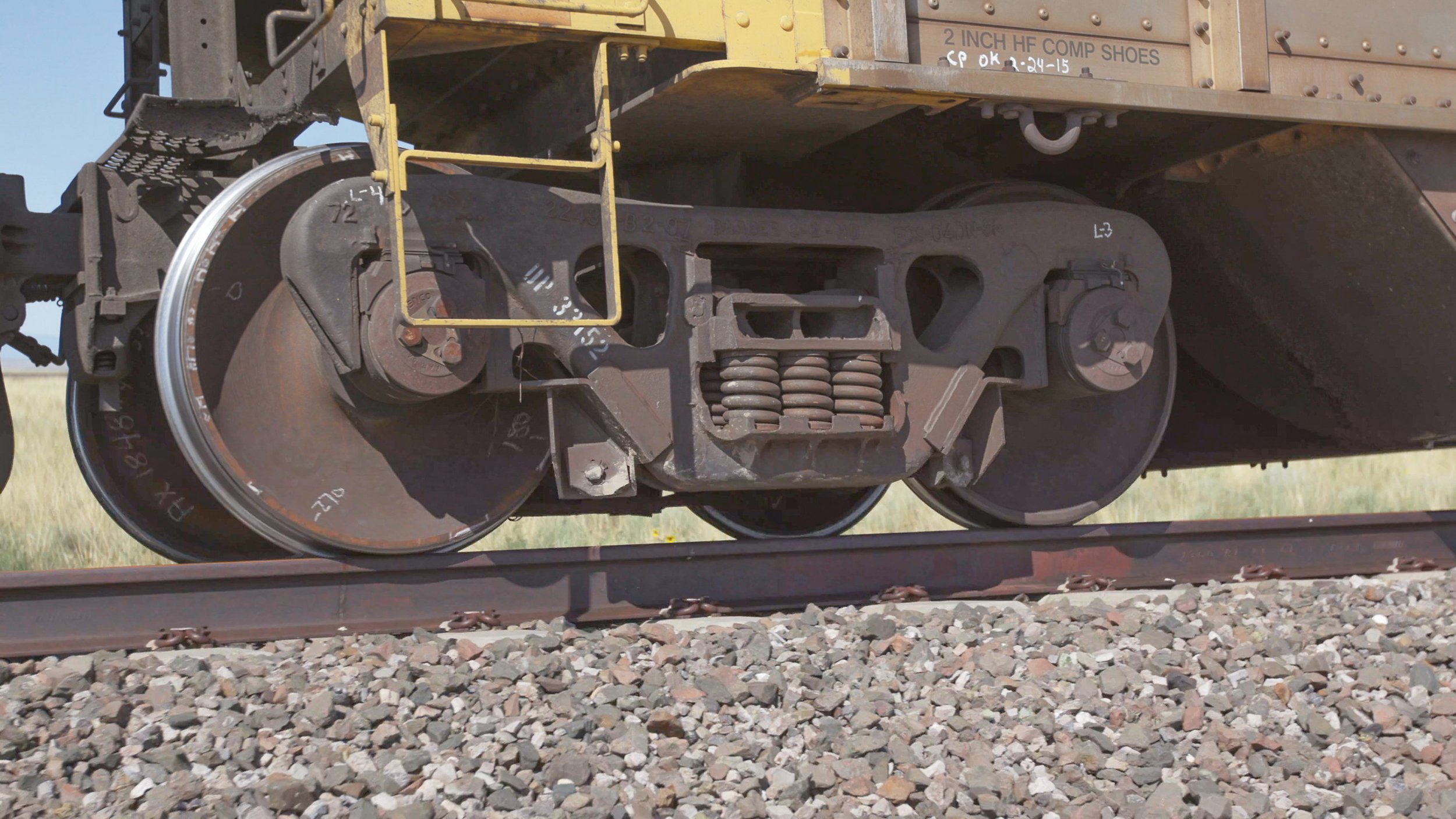 Close-up of a railway wheel and coupling mechanism on a train, with gravel on the tracks in the foreground.