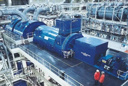 Industrial facility with large blue machinery, pipes, and two workers wearing red jackets.