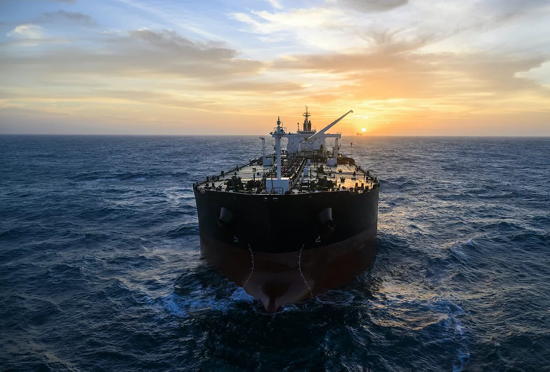 A large cargo ship sailing in the open ocean at sunset, with a partly cloudy sky and the sun near the horizon.