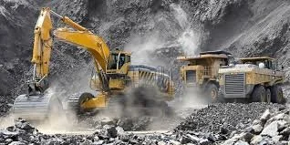 A yellow excavator digging into rocky terrain with dust and debris flying, and a large dump truck nearby.