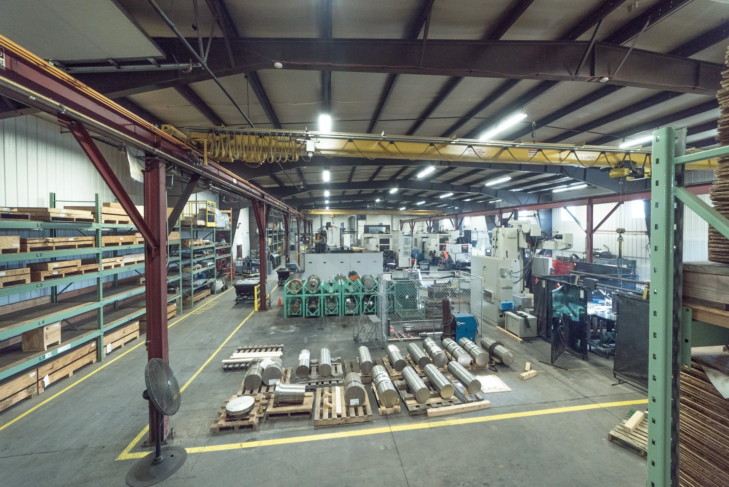 Interior view of a manufacturing warehouse with shelves of materials, industrial equipment, and metal parts on pallets.