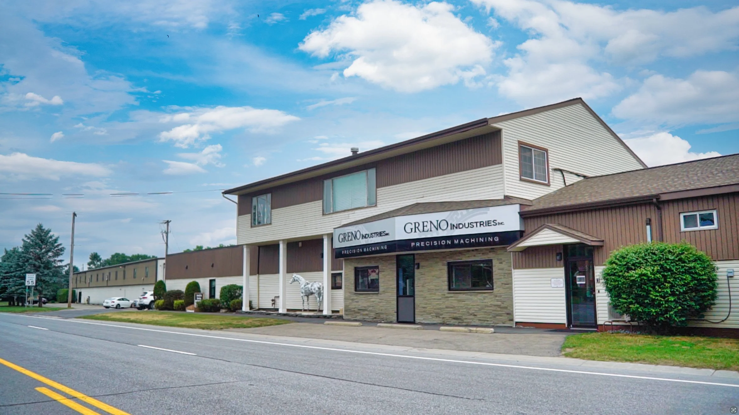 A commercial building with a sign reading 'Greno Industries Inc.' and 'Precision Machining' on a cloudy day. The building has two stories, with a small landscaped area featuring a white horse statue and some bushes. There are cars parked along the street in front of the building.
