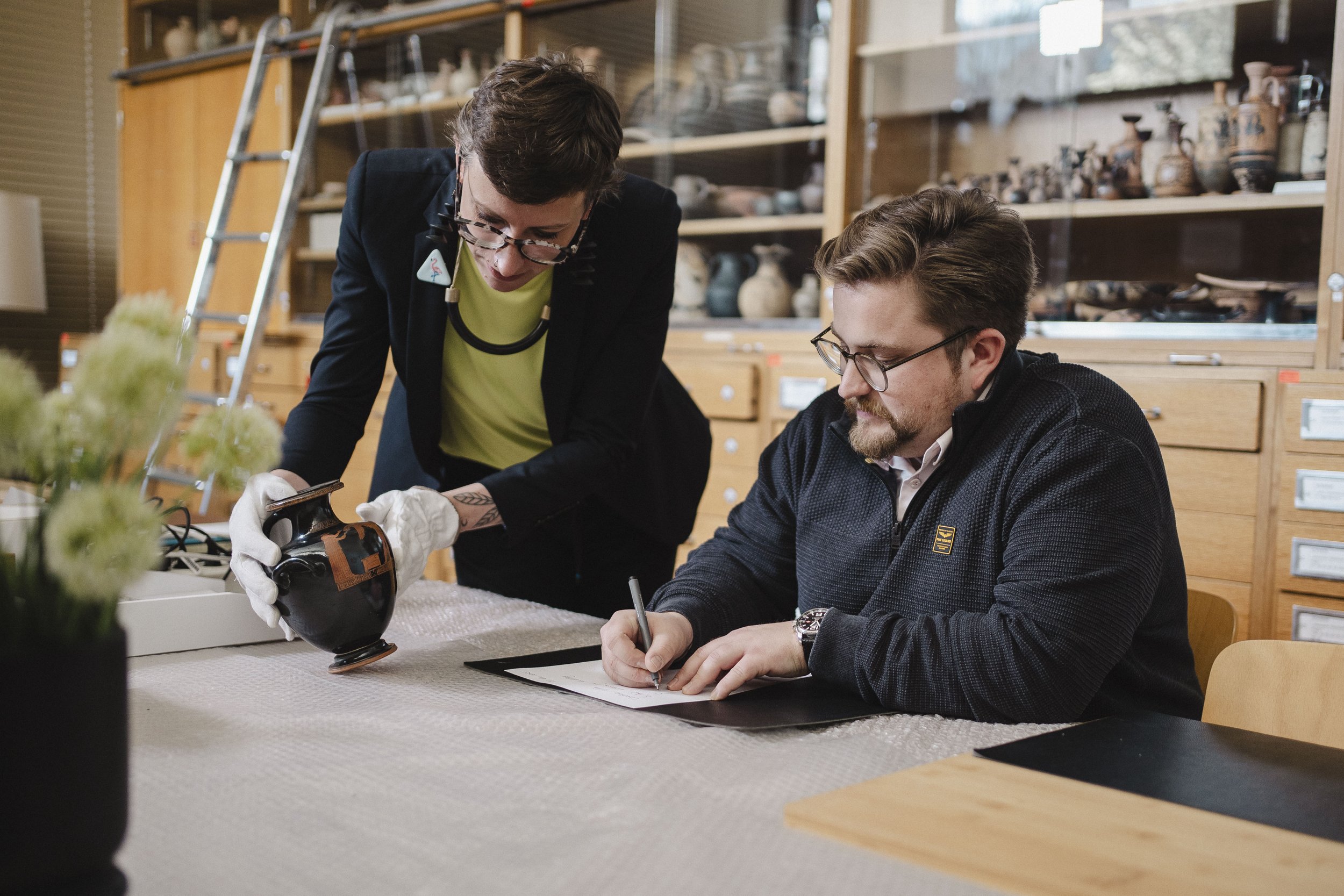 Polly Lohmann und Sascha Marschner, Vorstand der Novus Stiftung, bei der gemeinsamen Arbeit in der Antikensammlung Heidelberg: Sie inspizieren eine antike Vase und machen sich Notizen – Einblick in archäologische Forschung und Stiftungsarbeit.