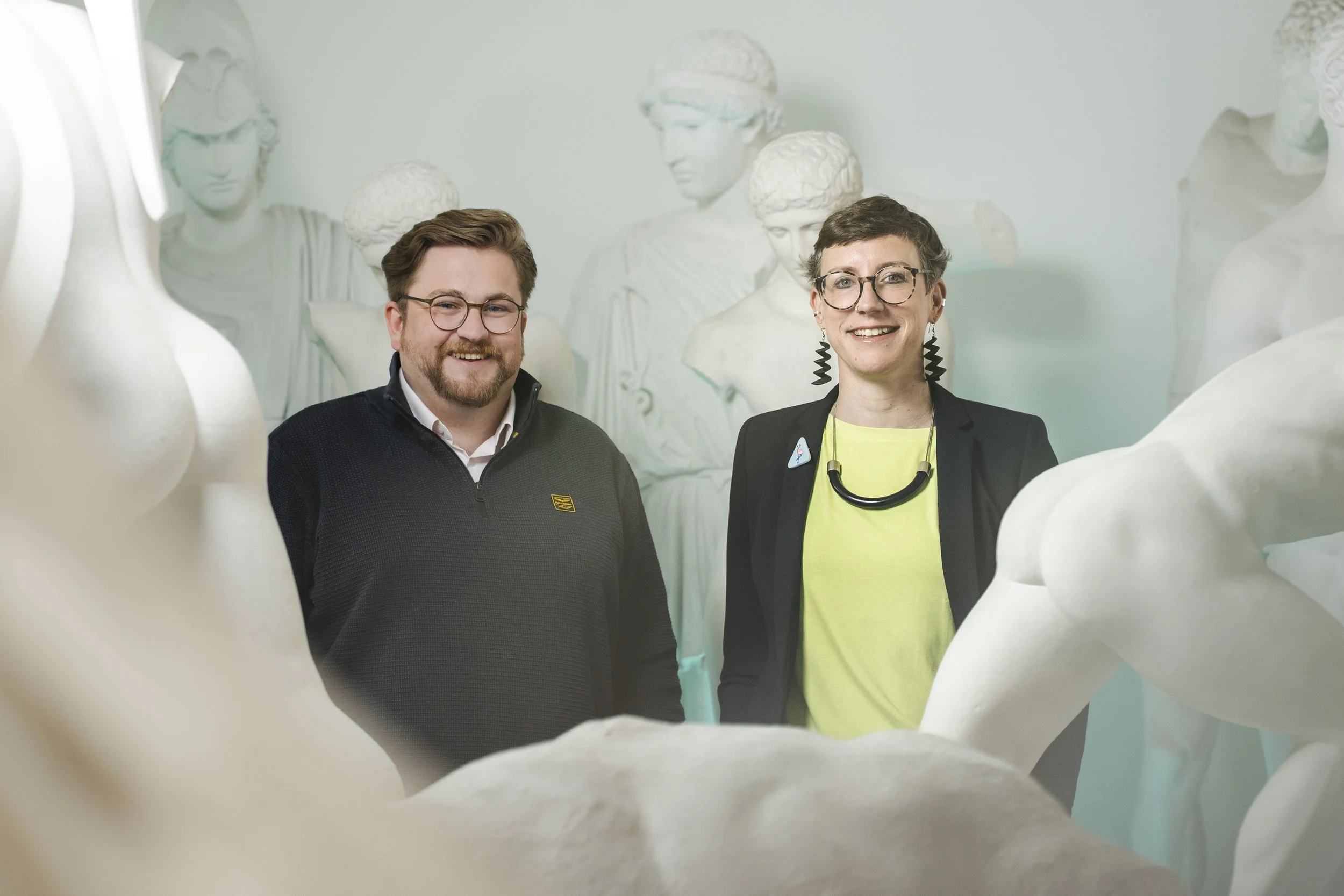 Gemeinsames Porträt von Sascha Marschner und Dr. Polly Lohmann, Vorstand der Novus Stiftung, freundlich und lachend zwischen Gipsabgüssen antiker Statuen in der Antikensammlung der Universität Heidelberg.