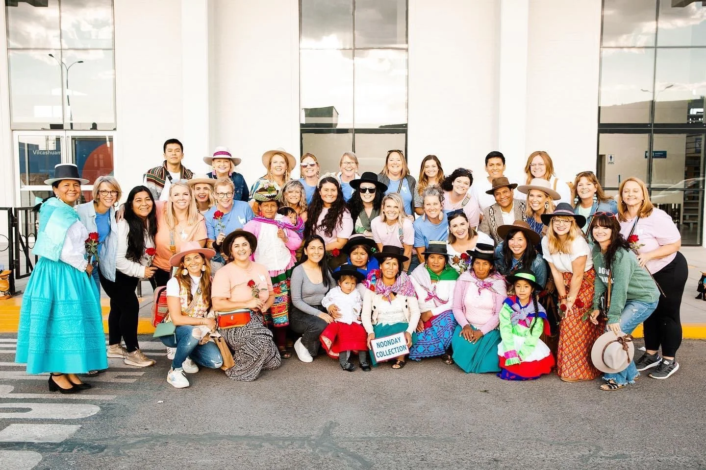 Group of diverse women and children in colorful traditional and modern clothing, posing together outside a modern building, some holding roses and a sign that reads 'NOONDAY COLLECTION'.