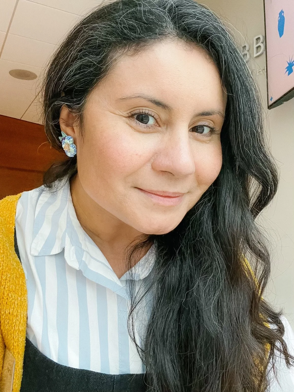 A woman with long black hair wearing blue floral earrings, a white and blue striped shirt, and a yellow cardigan, smiling indoors.