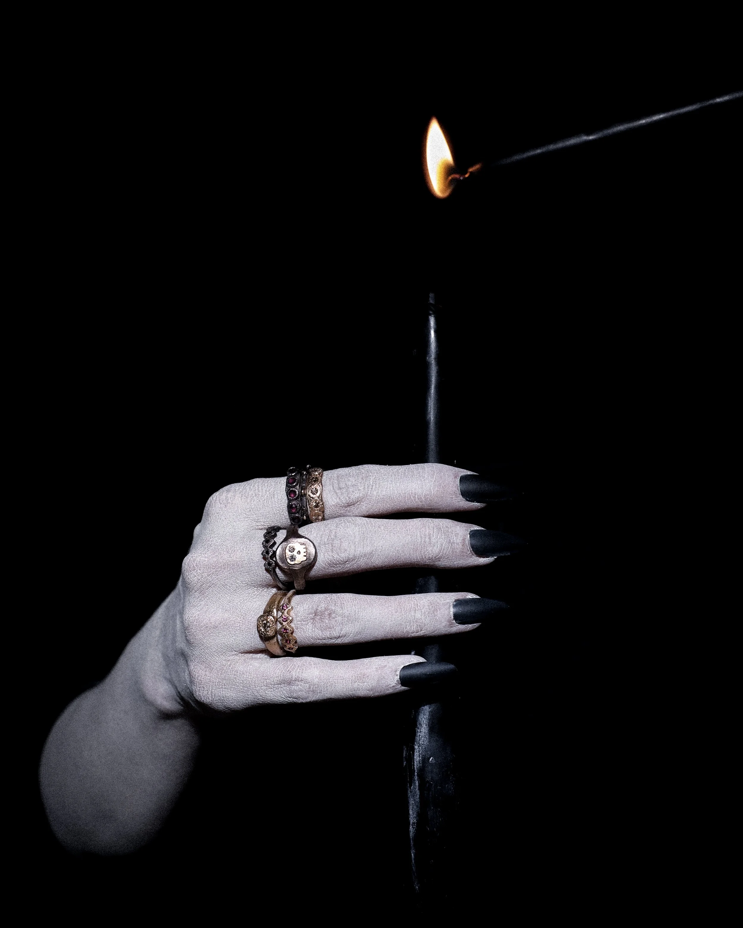 A hand wearing rings and a bracelet, holding a matchstick with a lit flame. The background is black.