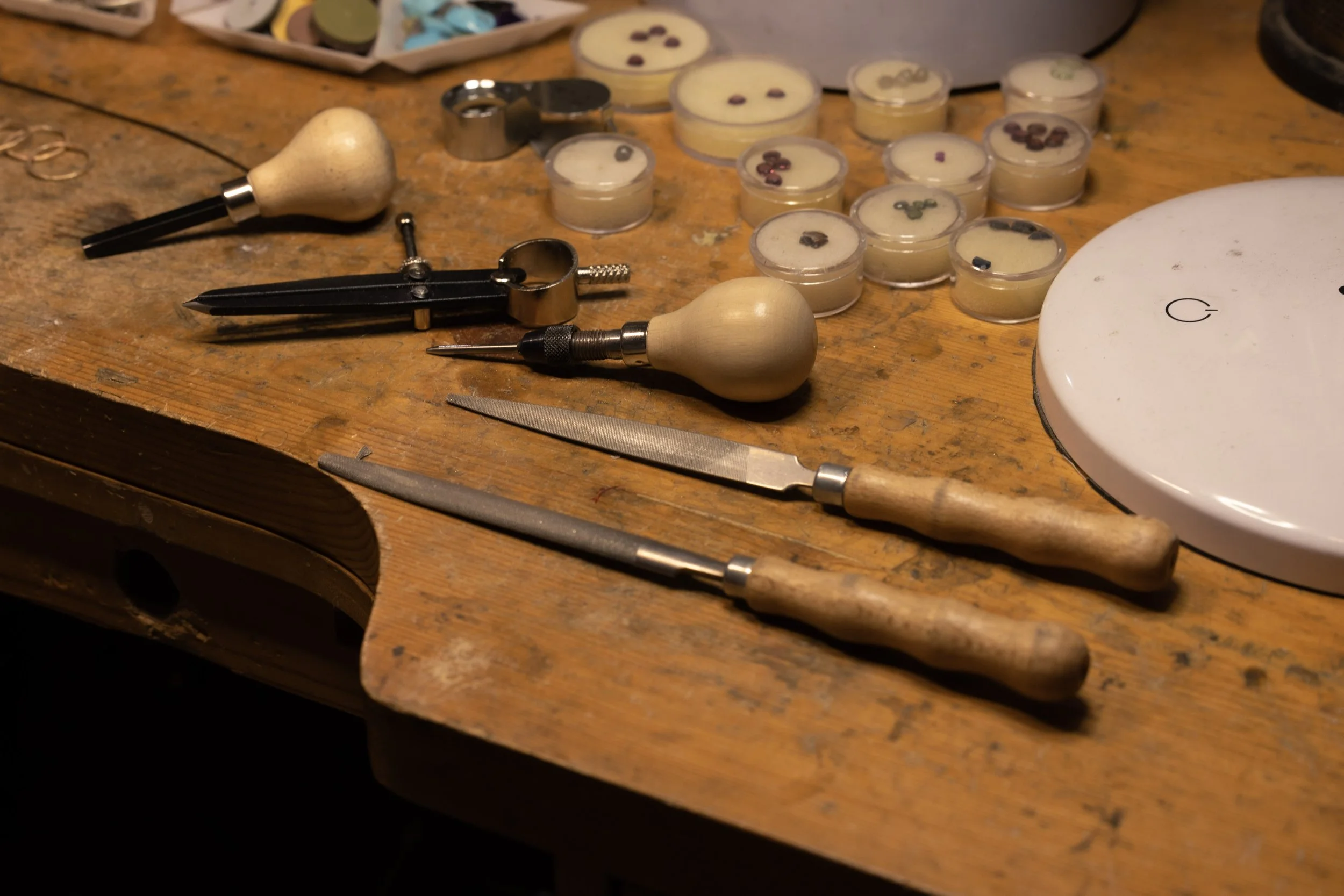 A wooden workbench with jewelry-making tools, including small files, screwdrivers, and hobby knives, along with small containers of beads or gemstones in the background.